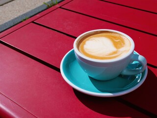 Latte or cappuccino in blue cup on red outdoor table – cobblestone street, scooter, parked cars, greenery, urban café vibe, coffee close-up, relaxing city moment, vibrant color contrast