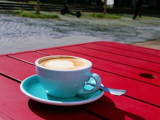 Latte or cappuccino in blue cup on red outdoor table – cobblestone street, scooter, parked cars, greenery, urban café vibe, coffee close-up, relaxing city moment, vibrant color contrast