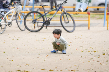 A young boy is playing in the park with a bicycle. The boy is crouching down and looking at something on the ground. The scene is peaceful and calm, with the boy enjoying his time outdoors © Masakazu Tokashiki
