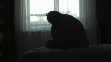 Silhouette of a millennial woman sitting on a bed in the dark bedroom, depression, pain. Portrait of Unhappy sad woman sitting against window and thinking about her life