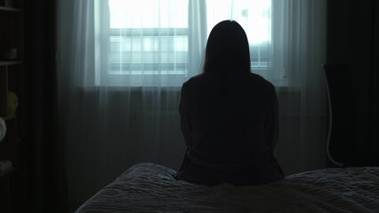 Back view Silhouette of a millennial woman sitting on a bed in the dark bedroom, depression, pain. Portrait of Unhappy sad woman sitting against window and thinking about her life