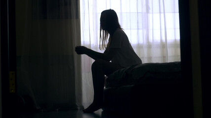 Silhouette of unrecognized woman sitting on a bed in the dark bedroom, depression, pain. Portrait of Unhappy sad woman sitting against window and thinking about her life