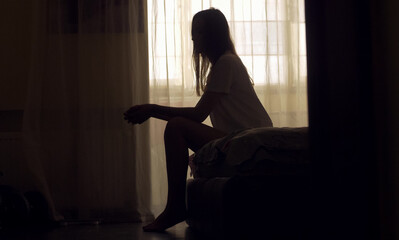 A woman sits alone on the edge of a bed in an empty room, her silhouette framed by soft light. She appears lost in thought, emotionally overwhelmed, facing a personal mental health crisis.
