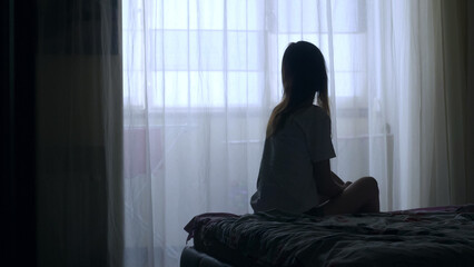 Silhouette of unrecognized woman sitting on a bed in the dark bedroom, depression, pain. Portrait of Unhappy sad woman sitting against window and thinking about her life