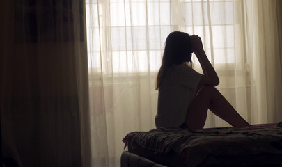 A woman sits alone on the edge of a bed in an empty room, her silhouette framed by soft light. She appears lost in thought, emotionally overwhelmed, facing a personal mental health crisis.
