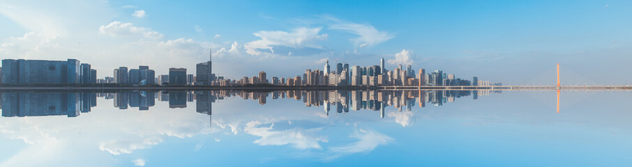 Urban Skyline Reflection on Water with Blue Sky and Clouds Panoramic View Featuring Buildings and Bridge Architecture Mirrored on the Calm Surface