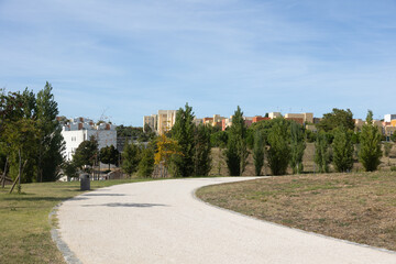 Carnide, Lisbon, Portugal - Curved walking path in Parque Verde surrounded by trees and greenery, with residential buildings in the background under a clear blue sky.