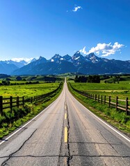 A long, straight asphalt road vanishes towards majestic snow-capped mountains under a vibrant blue sky, flanked by verdant fields and wooden fences
