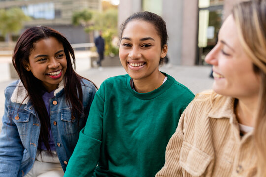 Group of three diverse student friends is sitting on a bench at the university campus, talking and laughing together. Latin American girl smile at camera. Friendship and youth community concept - Powered by Adobe