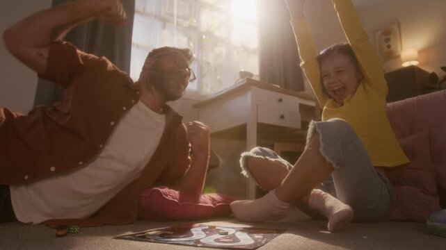 Low angle cinematic shot of cute young girl blowing on dice for luck while playing board game with father and cheering celebrating victory