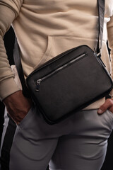 man dressed in a beige hoodie showcases a black bag emphasizing his casual style. He stands in front of a dark background adding depth to the scene.