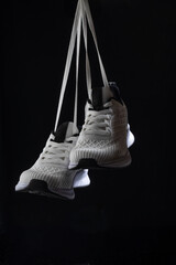 White sneakers dangle from their laces in front of a stark black background emphasizing the sleek design and modern aesthetics of the footwear. A minimalist setup highlights the shoes well.