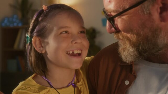 Close up shot of cute young girl embracing father and smiling happily sharing tender moment together at home lit by soft sunlight