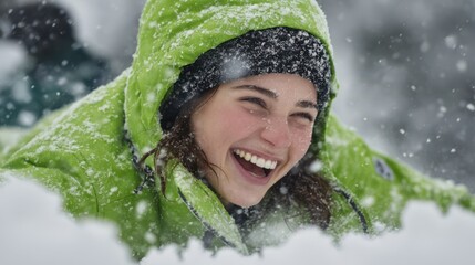 Close-up of laughing young woman in green winter jacket covered with snowflakes, enjoying playful outdoor moment during cold snowy day in winter season