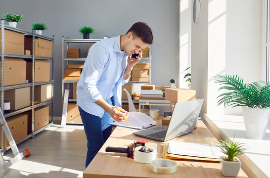 Young business man phone talk, laptop working online, checking box item title, price. Individual seller starting small business in warehouse storing area, marketplace manager receives ordered parcel