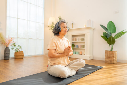 Happy Asian senior woman meditating in lotus pose with sound therapy on headphones at home. Healthy elderly woman enjoy wellness healthcare lifestyle relaxing, meditation yoga exercise in living room. - Powered by Adobe