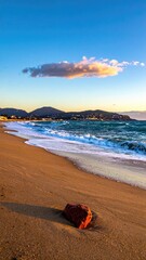 Serene coastal scene at sunset; gentle waves lap a sandy beach, a single reddish rock rests in the foreground under a clear sky