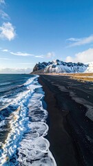 Aerial view of a black sand beach meeting a wave-lapped ocean under a clear blue sky, snow-capped mountains in the background