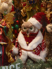 Festive owl wearing Santa outfit sitting among nutcrackers, angels, baubles, creating playful holiday scene with sparkling tinsel background
