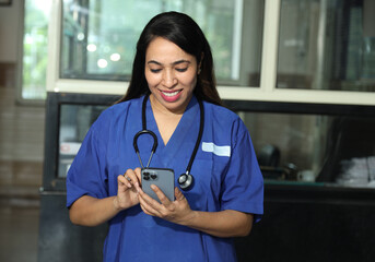 Indian happy female nurse or doctor using smart phone while standing in hospital. Beautiful young nurse woman wearing uniform and stethoscope. Concept Healthcare