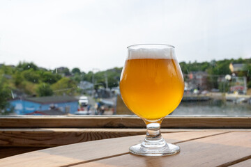 A tulip-shaped clear beer glass filled with a cold blonde ale on a wooden table. The booze has a layer of froth on top of the liquor. The table is dark wood, and there's a window in the background.