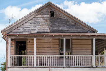 The facade of a small, worn, and weathered cottage. The wooden building has a peaked roof, broken windows, peeling paint, and a covered porch. The background is a bright blue sky with white clouds.
