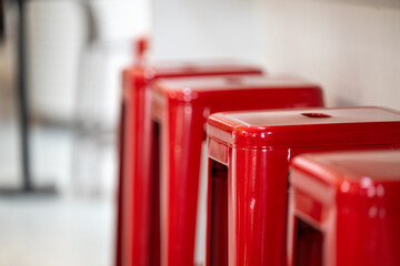 A row of bright red stackable metal bar chairs against a white interior wall. The square retro-style chairs have handle openings in the middle of the seat. The chairs are bar height. 