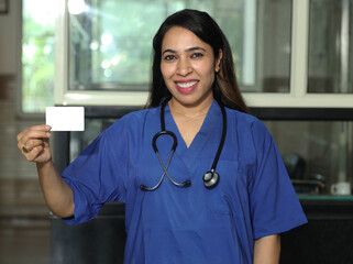 Indian happy female nurse showing business card. while standing in hospital. Beautiful young nurse woman wearing uniform and stethoscope. Concept Healthcare