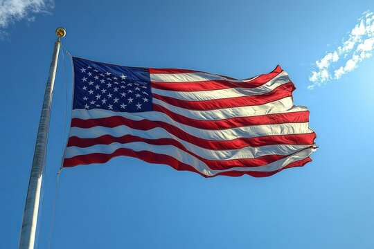 American flag waving strongly on a tall flagpole against a clear blue sky with a few scattered clouds