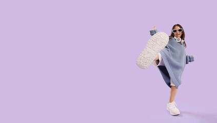 Cute little Asian girl in oversized sweater and with headphones dancing on lilac background with space for text
