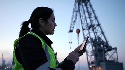 Female worker using smartphone with crane at the port during twilight hours - Powered by Adobe