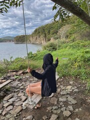Woman in hooded black fleece jacket sitting on the diy swing on the sea shore