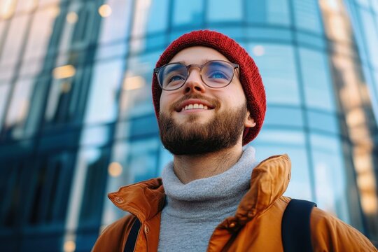 young man wearing glasses and a red knit hat smiling outdoors in front of a modern glass building on a cool day