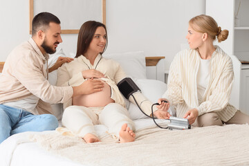Young happy pregnant woman with her husband and doula measuring blood pressure on bed in bedroom