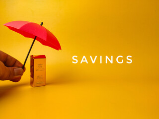 The image depicts a hand holding a red umbrella over a gold bar, with the word SAVINGS on a yellow background