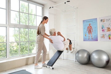 Fototapeta premium Female physical therapist assisting a senior man with abdominal exercises on an incline bench in a rehabilitation center