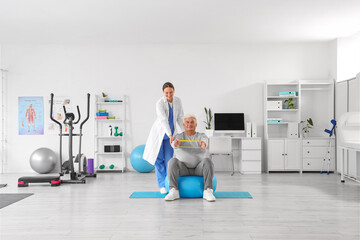 Female therapist and senior man with stretching band on fitball in rehabilitation center