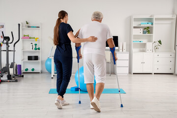 Female therapist and senior man with crutches walking in rehabilitation center, back view