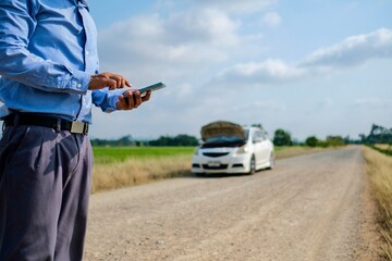 A man stands using his mobile phone to call for help because his car broke down on a dirt road far from his house.