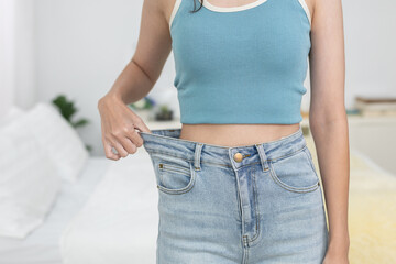 Slim woman wearing loose jeans showing weight loss and body transformation, symbolizing fitness,...