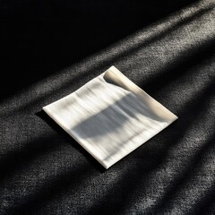 A square, off-white object, draped, on dark surface, sunlit