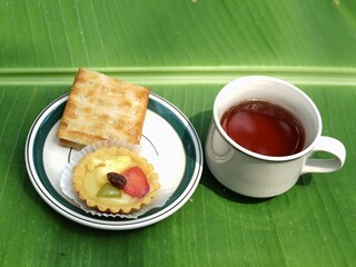 A white mug of hot tea sits next to a plate holding a fruit tart and cracker, all set against a vibrant green banana leaf background for a tropical tea break.