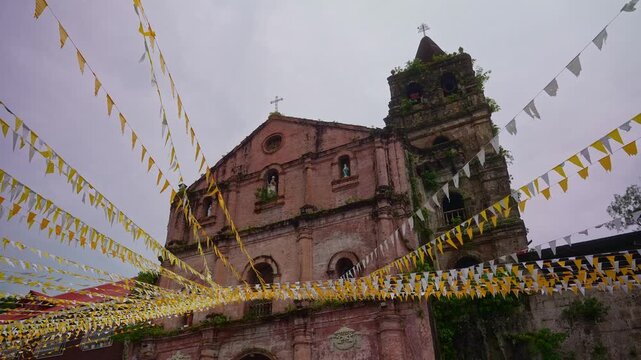 A low angle fixed shot of Minor Basilica and Parish of St. Gregory the Great, adorned with yellow and white banderitas swaying in the wind, under an overcast in Majayjay, Laguna Philippines