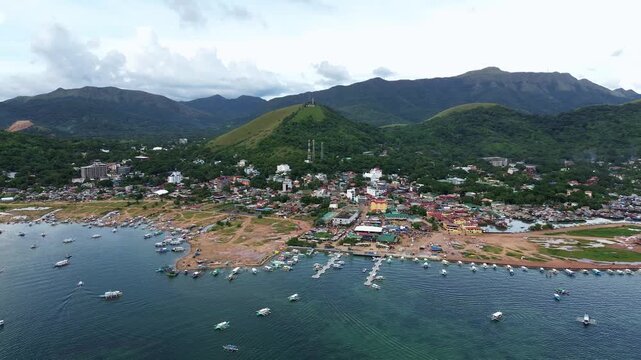 Wide aerial view over Coron town waterfront and moored boats with Mount Tapyas hill and surrounding green ridges under cloudy daylight in Palawan