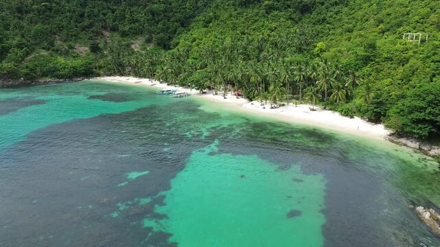 Aerial view above turquoise water and a palm lined white sand beach near Port Barton with reef patches and forested slopes under daylight