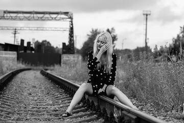 Young woman with long blonde hair, wearing a polka dot dress, sitting on railway tracks surrounded...