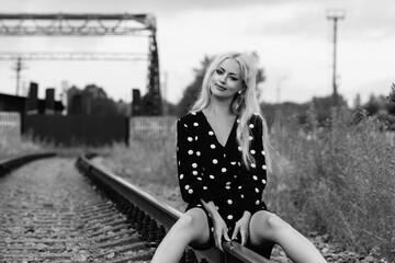 Young woman with long blonde hair, wearing a polka dot dress, sitting on railway tracks surrounded by overgrown grass, evoking a sense of freedom and adventure in a monochrome setting