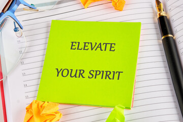 A bright green sticky note displays the phrase elevate your spirit, surrounded by colorful paper scraps, glasses, and a pen on a lined notebook. This workspace invites creativity and inspiration