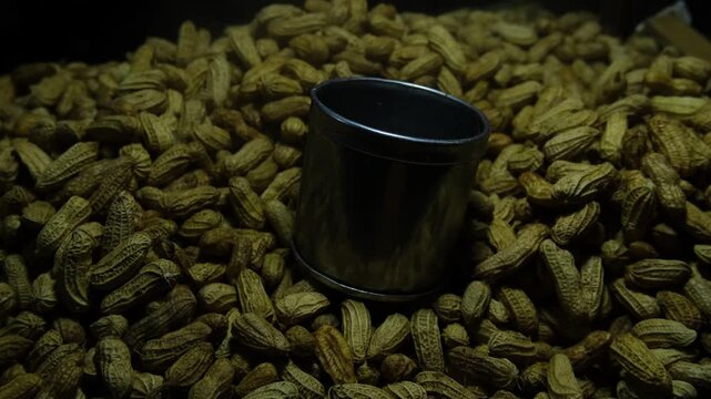 Close-up footage of a shiny metal measuring cup resting on a heap of dry, unshelled peanuts.