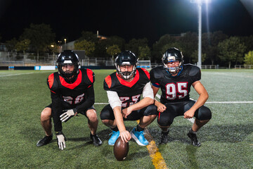 American football players in uniform and helmets crouching on a green turf field at night, ready for competition